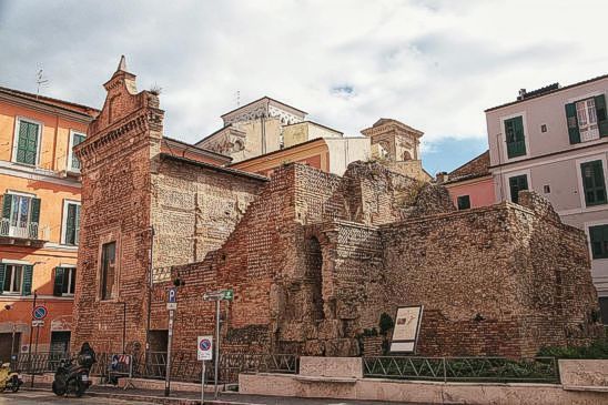 Chieti, antica Teate Marrucinorum, sorge a 330 metri sul livello del mare, su una collina che offre un panorama spettacolare che spazia dalla Maiella al Morrone, fino alla catena del Gran Sasso, i Monti della Laga e i Monti Gemelli. La città domina sia la Valle della Pescara a nord-ovest che quella dell'Alento a sud-est, abbracciando in questo modo un vasto territorio che giunge fino al mare Adriatico, in direzione di Pescara e Francavilla al Mare. Teate fu un importante centro del popolo italico dei Marrucini, ed è stata definita la "res publica" della loro comunità. Durante la Seconda Guerra Punica, la città fu alleata di Roma, mentre si oppose a essa durante la Guerra Sociale, aderendo alla Lega Italica di Corfinio. In seguito, divenne un centro di rilievo all'interno della Regio IV di Augusto, tra il I secolo a.C. e il II secolo d.C., ricoprendo un ruolo di fondamentale importanza nel panorama politico e culturale dell'epoca. L'evoluzione di Teate è testimoniata dai numerosi monumenti che ancora oggi caratterizzano la città. La crescita della città fu particolarmente significativa dopo la Guerra Sociale, quando importanti famiglie locali, come i Pollioni e i Vezii, contribuirono alla realizzazione di edifici pubblici e monumenti. Templi, un teatro romano, un anfiteatro e un ampio complesso termale sono alcuni dei principali esempi che testimoniano il massimo splendore della città. Questi edifici sono ora custoditi nel Museo Nazionale Archeologico alla Villa Comunale e nel Museo della città alla Civitella, insieme all'anfiteatro che è stato recentemente recuperato. I reperti archeologici attestano che la collina di Chieti è stata abitata sin dall'età del ferro, con un primo nucleo di insediamento che risale al 1500 a.C., mentre la tradizione locale fa risalire la fondazione della città al 1181 a.C., con leggende che la collegano a figure mitologiche come Achille e alla dea Teti. Il Teatro Romano di Teate, che risale probabilmente al I secolo d.C., è uno degli esempi più importanti dell'architettura della città antica. Il teatro, di forma circolare e con una capienza di circa 5.000 spettatori, conserva ancora parte della sua cavea, che misura oltre 80 metri di diametro. La struttura, che sfruttava in parte il pendio naturale della collina, era costruita con calcestruzzo e rivestita con pietra e mattoni disposti secondo la tecnica dell'opus reticulatum. L'ingresso principale al teatro era accessibile tramite una ripida salita, che portava i visitatori a un corridoio che si collegava alla cavea. Questo impianto testimonia non solo l'importanza del teatro come luogo di intrattenimento, ma anche la grandezza e la vitalità della città romana di Teate. Accanto al teatro, sono stati riportati alla luce i tempietti romani, noti anche come tempietti di S. Paolo. Scoperti nel 1935, questi monumenti sono costituiti da tre tempietti affiancati, di cui i primi due presentano una cella, un pronao e una cripta, mentre il terzo, più piccolo, è privo di pronao e presenta solo la cella e la cripta. La struttura muraria dei templi, costruita in calcestruzzo e rivestita in opus reticulatum, riflette il periodo di massimo splendore della città, che corrisponde al regno dell'imperatore Claudio. I resti di questi templi, insieme ai numerosi reperti rinvenuti, tra cui monete, lapidi e frammenti scultorei, dimostrano l'importanza religiosa della città e il suo continuo sviluppo nel corso dei secoli. La presenza di un quarto tempio romano nelle vicinanze, con fondamenta in tufo e pietra calcarea, conferma ulteriormente il ruolo centrale di Chieti nell'ambito religioso e civile dell'Italia antica.