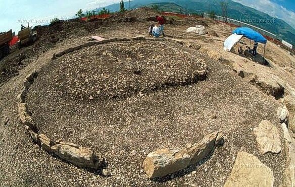 Il sito archeologico di Bazzano, situato nella frazione omonima de L'Aquila, è una delle necropoli più importanti e vaste della regione Abruzzo. Scoperta alla fine degli anni '80 e oggetto di scavi sistematici dal 1992, la necropoli ha rivelato oltre 1600 tombe che coprono un ampio arco temporale, dal IX secolo a.C. fino al I secolo d.C. L’area degli scavi, che si trova sotto l'attuale nucleo industriale di Bazzano, è stata indagata principalmente a causa dei lavori di costruzione degli edifici industriali, che hanno richiesto sondaggi archeologici preventivi. La necropoli non sembra essere associata a un insediamento specifico, ma rappresenta un importante centro funerario per diverse comunità che abitavano la regione nel corso dei secoli. Le prime testimonianze rinvenute nella necropoli di Bazzano risalgono alla prima età del Ferro, con sepolture a tumulo che conservano corredi funerari significativi, come vasi in ceramica, fibule in bronzo e cinturoni ornamentali. Le tombe di questa fase sono visibili in superficie e spesso danneggiate dalle lavorazioni agricole, ma alcune, come la tomba 690, sono ben conservate e offrono dettagli preziosi sulla cultura funeraria del periodo. In particolare, la tomba 690, che conteneva il corpo di un bambino, è un esempio emblematico delle sepolture a tumulo, che avevano una struttura a grande diametro, arricchita da stele di pietra. Queste tombe riflettono una società che già a partire dalla prima età del Ferro aveva sviluppato una cultura materiale complessa. L'area funeraria di Bazzano ha restituito anche sepolture databili alla metà del VIII secolo a.C., periodo in cui si osserva una particolare organizzazione delle tombe. Le sepolture di questa fase sono distribuite in modo ordinato e delimitate da una palizzata lignea, un elemento che è stato successivamente riscontrato anche in altre necropoli vestine, come quella di Cinturelli a Caporciano. Tra i ritrovamenti più significativi di questo periodo vi è la tomba 953, che ha restituito il cosiddetto "Principe di Bazzano", un individuo adulto sepolto con un ricco corredo di armi e vasellame in bronzo. L'influenza culturale dei Pretuzi, popolazione che abitava l'attuale provincia di Teramo, è evidente in alcuni oggetti, come il calice in impasto bucchero ritrovato nella stessa tomba, che testimonia gli scambi tra le diverse tribù italiche. Nel corso dei secoli successivi, la necropoli di Bazzano ha continuato a essere un luogo di sepoltura per le comunità locali, come testimoniato dalle tombe ellenistiche databili tra il IV e il I secolo a.C. Tra le sepolture di questo periodo si trovano corredi di grande pregio, come quello della tomba 833, che include ornamenti in bronzo, argento e pasta vitrea, con pendenti di origine cartaginese. In aggiunta alle tombe a fossa tradizionali, sono state rinvenute sepolture particolari, come quelle in grandi casse lignee con cerniere angolari in ferro, e tombe a camera, all’interno delle quali sono stati trovati letti funerari decorati con appliques in osso. La necropoli di Bazzano si conclude con una testimonianza della fase imperiale-romana: un asse stradale rivestito di basoli litici, su cui sorse una mansio, struttura che ospitava i viandanti e che rimase in funzione fino all’età alto-medievale.
