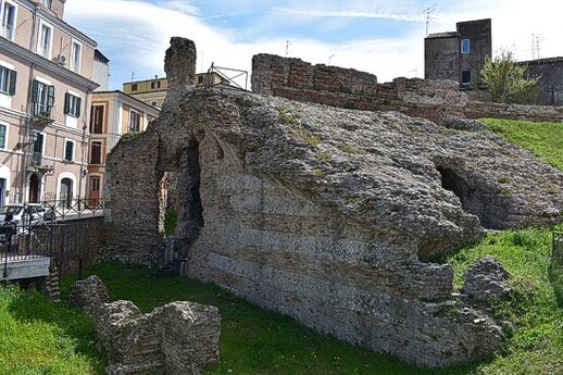 Il Teatro Romano di Chieti, costruito nel I secolo d.C., è uno degli esempi più significativi dell’architettura teatrale romana. Situato in una zona strategica della città, nei pressi di uno degli ingressi principali, l’edificio venne eretto su un terreno che in precedenza ospitava abitazioni private tardo-repubblicane. La cavea del teatro, che aveva un diametro di circa 80 metri, si adattava perfettamente al pendio collinare, permettendo così una visibilità ottimale per il pubblico. Lo sfondo scenico del teatro era incorniciato dal suggestivo panorama della Maiella, rendendo ogni spettacolo ancora più spettacolare. Nel corso dei decenni successivi alla sua costruzione, il teatro subì vari interventi di ristrutturazione. Tra questi, l’innalzamento delle gradinate della cavea, fatto per aumentare la capacità del pubblico, suggerisce un’espansione della città e un crescente interesse per gli spettacoli pubblici. Questi rifacimenti testimoniano l’importanza che il teatro ricopriva nella vita culturale della città, diventando uno dei principali luoghi di incontro e svago per la comunità teatina. L’edificio, con la sua struttura imponente e il suo utilizzo sociale, rispecchiava la prosperità e la vivacità della città romana di Teate. Tuttavia, con il passare dei secoli e il cambiamento delle dinamiche urbane, il teatro subì un progressivo degrado. A causa dei numerosi smantellamenti e delle modifiche apportate al sito, molte delle sue strutture furono distrutte o sepolte. L’edificio fu infine inglobato nell’espansione della città moderna e rimase nascosto fino alla sua riscoperta a metà dell’Ottocento, quando alcuni lavori edilizi rivelarono la sua esistenza. Questo ritrovamento non solo ha permesso di riportare alla luce l’antico teatro, ma ha anche sollevato l’interesse per il suo studio e la sua conservazione. Oggi, gli scavi archeologici condotti fin dagli anni ’30 hanno permesso di recuperare e preservare la porzione orientale della cavea e alcune strutture di accesso al teatro. Questi resti offrono uno scorcio prezioso sulla grandiosità dell’edificio originale, permettendo ai visitatori di apprezzare l’ingegneria romana e il ruolo che il teatro svolgeva nella vita quotidiana dell’antica Chieti. Il Teatro Romano di Chieti, nonostante il passare del tempo e i danni subiti, continua a rappresentare una delle principali attrazioni archeologiche della città, testimoniando la sua rilevanza nel panorama culturale e storico dell’antichità.