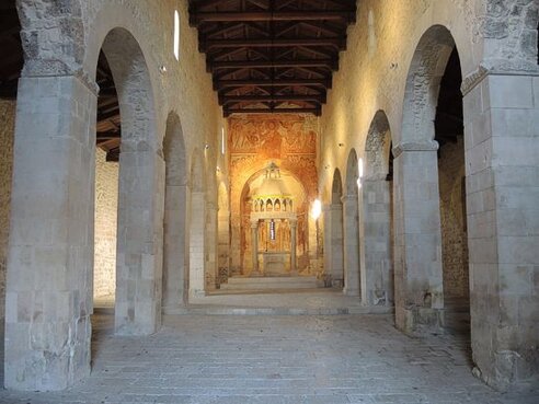 I luoghi sacri in Abruzzo: La chiesa di San Pietro “Ad Oratorium” (Interno). La chiesa di San Pietro ad Oratorium, situata ai piedi di Capestrano, lungo il corso del fiume Tirino, è un luogo di culto che affascina per la sua posizione isolata e immersa nella natura. Circondata da un boschetto di salici e pioppi, la chiesa sembra nascere dall'ambiente stesso, quasi come se fosse stata pensata per fondersi con il paesaggio circostante. La sua facciata semplice e rustica, caratterizzata dalle tre absidi semicircolari in pietra spoglia, trasmette un senso di intimità e silenzio, invitando i pellegrini a una sosta meditativa lontano dal trambusto del mondo. Nel cuore di questo luogo sacro, l'iscrizione enigmaticamente incisa nel concio rovescio della facciata richiama l'attenzione con il misterioso quadrato magico che recita “sator arepo tenet opera rotas”. Questa antica scritta, che sembra legare la chiesa a tradizioni e simbolismi lontani, diventa una sorta di porta simbolica per accedere al mistero di San Pietro ad Oratorium. L'architrave del portale, con l'indicazione della fondazione della chiesa da parte del re Desiderio, che nel 1100 fu rinnovata, unisce il culto religioso con la storia e la memoria collettiva. All'interno della chiesa, l'atmosfera è altrettanto suggestiva. Le possenti colonne che dividono lo spazio in tre navate creano un senso di grandiosità e maestosità, mentre il ciborio duecentesco, finemente scolpito, accentua la solennità del luogo. La sobrietà delle linee e dei dettagli, tipica dello stile romanico, permette a chi vi entra di percepire un forte senso di spiritualità, dove ogni elemento sembra avere il compito di orientare lo sguardo verso il divino. Particolare valore è dato agli affreschi del XII secolo, che decorano l'abside centrale. Dipinti in ocra rossa, questi affreschi, pur nella loro semplicità cromatica, trasmettono un'intensità spirituale che accompagna il visitatore nella riflessione. La chiesa di San Pietro ad Oratorium, con la sua struttura maestosa e i suoi tesori artistici, è un luogo che racconta una storia millenaria di fede e devozione, testimoniando la continuità del culto cristiano in Abruzzo e la sacralità dei luoghi che lo accolgono.
