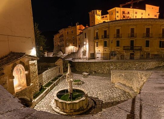 Fontecchio è un affascinante borgo medievale che si trova nel cuore della Valle Subequana, ai piedi del Monte Sirente, un luogo che ha mantenuto intatto il suo carattere storico. Il paese sembra essere uscito da un lontano passato, grazie soprattutto alla presenza della sua splendida fontana gotica, che è perfettamente integrata nel tessuto urbano originario. Questa fontana non solo arricchisce la bellezza del paese, ma rappresenta anche un simbolo della sua tradizione e della sua storia, testimoniando il legame con le epoche passate. Il borgo di Fontecchio è un classico esempio di paese fortificato sommitale, che si sviluppa su un’altura e viene protetto da due cerchie di mura concentriche. Questo assetto fortificato è ancora ben visibile nel centro storico, dove si possono ammirare le antiche mura e le porte che permettevano l'accesso alla cittadella. Oltre a questi elementi difensivi, il paese conserva palazzi fortificati e numerosi esempi di botteghe medievali, tra cui quelle con ingressi “zoppi”, che caratterizzano ancora oggi l’aspetto tipico del borgo. Un altro importante elemento storico di Fontecchio è la chiesa della Vittoria, situata nella parte più alta del paese. In passato conosciuta come la chiesa di San Pietro, questa struttura è stata costruita su un antico tempio dedicato a Quirino, divinità pagana. La chiesa testimonia la stratificazione storica del luogo, dove l'architettura medievale si sovrappone alle vestigia di epoche precedenti, creando una fusione unica di elementi culturali e religiosi. Fontecchio non è solo un borgo dalle bellezze architettoniche, ma un vero e proprio scrigno di storia, che racconta la vita medievale in Abruzzo. Ogni angolo del paese è impregnato di tradizioni e leggende che si intrecciano con la storia, facendo di questo piccolo borgo una meta imperdibile per chi desidera immergersi nel passato affascinante di questa regione. La sua posizione e la conservazione di elementi storici fanno di Fontecchio un luogo che custodisce gelosamente il fascino di un'epoca lontana.