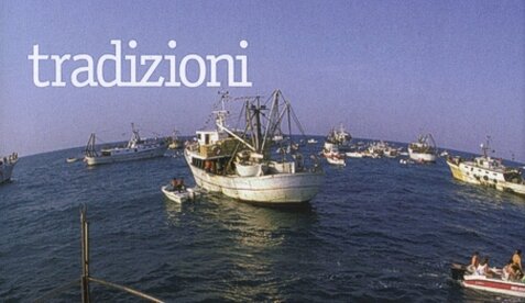 La festa della Madonna del Portosalvo con la processione dei motopesca a Giulianova Lido (Te). Nella seconda settimana del mese di agosto si tiene questa processione alquanto inusuale, che vede decine di imbarcazioni, addobbate a festa e stracolme di fedeli a bordo, solcare le acque antistanti la cittadina rivierasca. E sulla prima della flotta c’è la statua della Madonna del Portosalvo, protettrice dei pescatori. Qual che più colpisce i turisti, poco usi alle tradizioni della nostra terra, è l’osservare gli uomini di mare, rudi e cupi nell’immaginario collettivo, mentre con grande gusto e delicatezza infiorano di nastri e bandierine colorate i loro battelli. Il contrasto è netto, ma l’occasione lo merita: questo sfugge al turista. Il mare potrà anche plasmare a sua immagine l’aspetto di quelli che lo sfidano ogni giorno, per una vita, per poter campare, ma tira anche fuori il meglio dall’ animo umano. Oltre l'apparenza, il cuore degli uomini di mare è grande come quel blu senza fine che essi affrontano. Così pure grandi sono la loro fede e la loro riconoscenza verso Colei che li protegge, sempre vigile attimo dopo attimo, nel loro duro lavoro. Come in tutti i mestieri rischiosi, la religiosità profonda degli uomini semplici aiuta a vivere il pericolo giorno per giorno. Alla Madonna del Portosalvo, a Colei cioè che protegge gli uomini del mare, a Giulianova è dedicata una festa, una grande festa che ravviva di mille colori il bianco e la ruggine delle barche e il grigio delle banchine. Il legame della cittadina col mare è vecchio di cent’anni almeno. Al Lido tutto parla del mare: i pescatori che rammendano le infinite reti rosso mattone stese sul lungomare, le donne che vendono il pesce fresco sui loro curiosi carrettini, i vecchi marinai che hanno tante storie da raccontare quante sono le loro rughe, e ogni volta è un’ avventura diversa. È il porto il cuore vero della città. Ed è alle porte del ferragosto che il esso si anima di un fervore diverso, solo diverso, attenzione, non maggiore, perchè sulle banchine i passi corrono veloci in ogni momento, notte e giorno, e con ogni tempo. È il fervore della gente di mare che prepara la sua festa. Si cuocerà il pesce per offrirlo ai turisti e agli amici, si verserà il vino per brindare ai mille pericoli scampati e scacciare le ielle del domani, si suonerà la musica per gioire della gioia di tutti. E si giocherà, correndo come novelli cavalieri sul viscido palo di una cuccagna un po’ fuori dalle regole, proteso com’e dalla barca verso il mare e non verso il cielo. E si cadrà in mare mentre si corre verso il premio, così come si cade nella vita, e dal gran tuffo si tornerà sulla barca e di nuovo a sfidare il viscido che è sul cammino verso il traguardo. A decine ogni anno si sfidano i novelli cavalieri, a migliaia ridono dei loro folli voli in mare. Ma la festa non è solo questo. Certo si gioca, si mangia, si ride, ma se il turista gode la sagra paesana con spensieratezza, la gente di mare ha l’animo agitato di chi vive la vigilia. Solo un pensiero occupa la mente: la processione. E sarà la Domenica. È già dal mattino che tutta la gente di mare si affanna attorno al proprio battello per farlo più bello, più colorato, più festoso di tutti. Ma non è una gara tra barche, è una sfida a se stessi in onore di Colei che si va festeggiare. Tutti lo sanno. Lei li guarderà tutti mentre usciranno in mare per renderle omaggio e nessuno di loro vuol essere da meno quanto a colori e festoni. Quando il sole picchia ancora dritto sulla banchina d’asfalto infuocato, saranno neppure le tre del pomeriggio, l’ animo della gente di mare si accende dell’emozione per l’ ora cruciale che si avvicina. Non c’è caldo che tenga, si suda, si fatica, magari s’impreca, ma quando arriverà la processione tutto dovrà essere perfetto. Man mano che il sole scivola verso le colline, sale la tensione e sulle banchine cresce la folla: tutti vorrebbero salire sulle barche che stanno per uscire per rendere omaggio a Colei che protegge la gente di mare. Man mano che dalle vie della città sale il canto della processione, di pari passo cresce l’ emozione. Sulle barche ferve l’attività. Si formano gli equipaggi; mani dure di fatica aiutano le nere vecchine nel trasbordo, il dubbio inchioda per lunghi istanti i timorosi mentre osservano quanto siano terribilmente lunghi quei 3 palmi che separano, dal molo, la barca sulla quale vorrebbero salire. E se cado? Ma chi me l’ha fatto fare? Ma è un attimo. Solo un piccolo balzo, ed il morbido tonfo sul legno del ponte li accoglie a bordo. Nuvole nere fuggono veloci dai fumaioli, si mollano gli ormeggi, passeggeri dell’ ultim’ora guadagnano il loro posto in barca con balzi coraggiosi, infinite sono le tinte del blu che il cielo riflette nelle scie. Si va. Pian piano le acque del porto cominciano così a sorridere del riflesso di mille festoni colorati che sventolano sulle barche in festa. Come la folla che segue la Madonna, così le barche escono in fila indiana verso il mare aperto. Le grandi e le piccole, perchè non sono solo i grandi pescherecci a prendere parte alla festa; chiunque abbia un legno galleggiante si mette in scia. Tutti vogliono essere partecipi della festa che si va a cominciare. Fuori dal porto le scie si incrociano ormai a decine, motore al minimo, tensione al massimo. Ci sono tutti, manca solo la Barca, quella con la B maiuscola, quella che avrà quest’ anno l’ onore di portare a mare la statua. Esce? Perchè ancora non esce? I chierichetti hanno appena iniziato a calpestare la banchina. Nella processione l’ attesa allunga i metri man mano che i passi dei fedeli avanzano. A mo’ di novelle guide indiane, dalle barche ciascuno cerca di leggere nei movimenti della folla sul porto i segnali che lascino intuire dov’è arrivata la statua. Il vento porta lontano le note del pregare, ma qualcuno dal mare le ode, o lo crede, ed indica e grida: sono a metà! Sono alla curva! L’ alto muro della banchina copre la vista dal mare, solo gli alberi dei pescherecci emergono. Si muove, si muove! Allora ci siamo. È un segno inconfondibile, l’albero della Barca muove verso sud. Sta uscendo. Si va. E dietro di lei tutte le altre che la scortano. Tutti i motori assieme lanciano un ruggito fumoso, i solchi delle scie si fanno profondi, la brezza si alza per carezzare i passeggeri. All’apparire della Barca oltre la punta del porto la tensione ha un’esplosione: applausi e grida. La folla galleggiante in attesa inizia a disporsi quasi secondo un copione. La Barca fugge veloce dal porto facendosi largo tra le altre barche festanti. E loro si fanno da parte, e poi la scortano. Il grande gruppo è compatto, solo i veloci motoscafi sfrecciano facendo girotondo attorno ai più lenti pescherecci. Saranno cento, o forse di più. La confusione più ordinata che si possa immaginare. Si naviga fianco a fianco, pochi metri tra le fiancate. Gli agili barchini si divincolano tra i grandi con manovre da brivido, saltando di scia in scia. Il mare si infiamma verso terra del riflesso accecante di un sole ormai al tramonto. Saranno venti minuti che si naviga, trenta al massimo, ma a molti sembra di essere in mare da ore. Il turista che ha dimenticato di soffrire di mal di mare c’è sempre, ma la curiosità è tale che si stringono i denti e si tace. Ora il battere dei pistoni cala di intensità, il ponte vibra più leggero, solo un fil di fumo esce ormai dai fumaioli, la Barca vira leggera, le altre la imitano, ci siamo. È il momento più forte di una giornata che si ripete, unica, ogni anno. Giorni di preparazione, ore ed ore di tensione, tutto per dare vita ad un solo attimo di grande commozione. D’improvviso ci si stupisce come siano rimasti solo il battere dell’ onda sulle chiglie e il fruscio del vento tra le cime a rompere il silenzio. L’eco di una preghiera giunge lontana. È una corona verde d’alloro quella che vola verso il blu profondo. Dalla mano del parroco, dal cuore della gente di mare. È quello il loro modesto, semplice, vero omaggio ai compagni, agli amici, ai parenti che il grande blu ha voluto con se. Ma state certi, non c’è rabbia verso quel mare che li ha privati di tanto affetto, non risentimento verso quella distesa d’acqua che da sì loro da vivere, ma in cambio chiede tanto sacrificio. L’ animo della gente di mare è semplice. In loro c’è solo quel rispetto quasi religioso che l’uomo sente di dover portare alle cose più grandi di se. L’ urlo festoso delle sirene copre un grande applauso liberatorio. La tensione accumulata vola in un attimo al cielo come i gabbiani che fuggono alti, spaventati dalle scie dei razzi. Il rito è compiuto, la gente di mare ha reso omaggio ai suoi caduti, al mare e a Colei che li protegge. Si torna. Ora che la cerimonia è compiuta i motori sembrano andare più veloci, l’atmosfera è davvero quella della festa, stasera si ballerà e si canterà. I volti della gente di mare finalmente si distendono. La Barca non ha ancora finito di stringere gli ormeggi che già mani veloci sollevano la statua e, con mille attenzioni, la portano in trionfo. Sono questi uomini speciali: indosso la loro divisa bianca e blu, sul volto le rughe di mille e mille giorni in mare, nel cuore l’ orgoglio di essere Marinai, per sempre. Ora essi non calcano più i ponti delle navi da guerra, ma le loro emozioni tornano a navigare ogni volta che ai loro occhi appare il blu. Un tuono improvviso. I volti di tutti scattano al cielo mentre l’ aria esplode nella miriade di nuvole di fumo colorato di una grande batteria di fuochi d’artificio. Minuti che sembrano ore scandite dal ritmo crescente degli spari. Torna il silenzio. Le anime sulla banchina passano rapide dal tumulto del rombo al raccoglimento che la messa richiede. Una grande chiesa all’aperto. Il palco come altare, il molo come pavimento, le barche come pareti, le bici e gli scooter come banchi. Le grandi nuvole che avevano atteso finora pazienti, prendono veloci possesso del cielo, qualche barca sta ancora attraccando, ma le prime gocce di pioggia mirano già impietose i volti dei fedeli accalcati sulla banchina. Il tempo, si sà, non garantisce il sole neppure alla gente di mare nel giorno della loro festa. Ma la pioggia spaventa il turista, che corre sotto l’ombrello alla prima goccia; non loro. Non ci saranno nuvole, rovesci o marosi che li fermeranno.