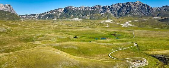 Campo Imperatore, conosciuto storicamente come "Campo Imperiale" per volontà di Federico II di Svevia, è un vasto altopiano di origine glaciale e carsico-alluvionale situato nella provincia dell’Aquila, nel cuore del massiccio del Gran Sasso e del Parco Nazionale del Gran Sasso e Monti della Laga. Con un'altitudine compresa tra i 1.500 e i 2.100 metri sul livello del mare, questo luogo offre paesaggi incontaminati e spettacolari, ideali per gli amanti del trekking e della natura selvaggia. Considerato il più vasto altopiano dell’Appennino e tra i più estesi d'Italia, Campo Imperatore si sviluppa lungo un asse nord-ovest/sud-est, con una lunghezza massima di 18 km e una larghezza di 8 km, per una superficie complessiva di circa 75 km². L'altitudine varia dai 1.460 metri della Val Voltigno fino ai 2.138 metri, raggiunti presso l’Osservatorio Astronomico. Il paesaggio è caratterizzato da ampie distese erbose, formazioni rocciose modellate dagli agenti atmosferici e una straordinaria varietà di flora e fauna che rendono ogni escursione un'esperienza unica e affascinante. Un percorso di trekking attraverso Campo Imperatore permette di immergersi completamente nella bellezza di questa prateria d’alta quota, dove il silenzio è interrotto solo dal soffiare del vento e dal richiamo degli animali selvatici. Lungo il cammino, una tappa imperdibile è la visita ai ruderi della chiesa di Santa Maria del Monte di Paganica, un luogo che conserva intatta un’atmosfera di sacralità e mistero. Tra le sue mura di pietra si avverte ancora la presenza della vita rurale del passato: l’eco delle voci, l’odore degli animali e la solidità dell’architettura che raccontano storie di un tempo lontano. Proseguendo lungo il sentiero, il paesaggio si apre su due piccoli specchi d'acqua, i laghetti di Passaneta e Racollo, che si rivelano come oasi di quiete immerse nella vastità dell’altopiano. Qui la natura si manifesta in tutta la sua autenticità, offrendo ristoro non solo agli escursionisti ma anche agli animali domestici e selvatici che abitano la zona. L’acqua limpida riflette il cielo e le montagne circostanti, creando scenari di rara bellezza che cambiano colore a seconda delle stagioni e della luce del giorno.