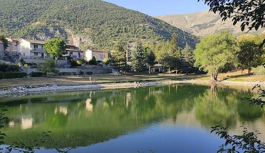 La Riserva Naturale del Monte Argatone si estende interamente nel territorio comunale di Villalago, in un paesaggio ricco di suggestioni naturali e spirituali. È suddivisa in due aree principali: una che comprende i bacini del fiume Sagittario e del Lago di San Domenico, l’altra che si sviluppa attorno al Lago Pio, incastonato tra i versanti del Monte Genzana e della Montagna Grande. Le acque smeraldine della riserva sono animate da piccole cascate e accolgono diverse specie di uccelli migratori. Tra gli elementi storici e architettonici spicca un antico mulino ad acqua, restaurato per valorizzarne la funzione e il fascino originario. Il Lago di San Domenico, situato nella parte alta della Valle del Sagittario, in località Prato Cardoso, è un luogo incantato ideale per momenti di relax e attività come la pesca. Le sue acque, intense nel verde smeraldo, sono ricche di fauna ittica, in particolare di trote fario. Un suggestivo ponte ad archi conduce all’unica spiaggetta del lago e all’Eremo di San Domenico, un piccolo santuario incastonato nella roccia, accessibile attraverso un portico impreziosito da una bifora decorata con vista sul lago. All’interno, la chiesa romanica conserva un altare neogotico di fine Ottocento, mentre sotto il portico esterno quattro dipinti del 1938, opera di Alfredo Gentile, narrano la vita del santo benedettino che qui visse attorno all’anno Mille. Il Lago Pio, di origine carsica, è alimentato da cunicoli e sorgenti sotterranee collegati al Lago di Scanno. La vegetazione circostante varia dalle rive ricche di salici e aceri, ai boschi di querce a bassa quota, fino alle faggete che si spingono verso le alture. La riserva ospita numerose specie floristiche di pregio, tra cui la genziana, orchidee rare, l’Ofride di Bertoloni e il giglio rosso, oltre a un’abbondante varietà di funghi. La zona è particolarmente apprezzata per il bearwatching, soprattutto tra agosto e settembre, quando la maturazione di particolari bacche attira l’orso marsicano. Con l'accompagnamento di guide esperte, è possibile osservarlo in sicurezza dai punti attrezzati predisposti dal Comune e dall’Ente Parco. Non mancano altre presenze faunistiche importanti come camosci, cervi, lupi, rapaci come il falco pellegrino e l’aquila reale, e uccelli acquatici come il germano reale, il merlo acquaiolo e la ballerina gialla. La riserva è attraversata da percorsi escursionistici, mentre la balneazione non è consentita. Nelle aree limitrofe si trovano altre importanti aree protette come la Riserva Naturale delle Gole del Sagittario, il Parco Nazionale d’Abruzzo, Lazio e Molise e la Riserva Naturale Monte Genzana.