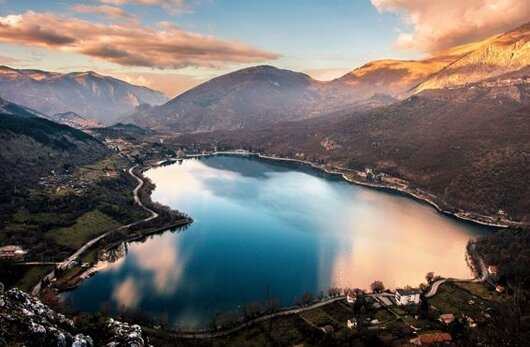 Il Lago di Scanno, incastonato tra le montagne dell’Abruzzo, è un gioiello naturale che deve la sua origine a una frana del Monte Genzana, che ha sbarrato la valle del Sagittario. Situato a 922 metri di altitudine, il lago si distende in una conca circondata da fitti boschi e dai borghi di Scanno e Villalago, offrendo un paesaggio di rara bellezza. Le acque, dal caratteristico colore verde smeraldo, riflettono le cime circostanti, creando un’atmosfera suggestiva che affascina visitatori e fotografi in ogni stagione. A est, la Riserva del Monte Genzana incornicia il bacino con le sue imponenti pareti rocciose, contribuendo al fascino selvaggio di questo angolo d’Abruzzo. Le acque del lago, che raggiungono una profondità massima di 36 metri, vengono alimentate dal fiume Tasso, mentre il deflusso avviene attraverso sorgenti sotterranee, mantenendo il livello idrico costante. Questo equilibrio naturale ha reso il Lago di Scanno una meta prediletta per gli amanti della natura e del turismo climatico. Le sue rive ospitano bagnanti in estate e sportivi appassionati di canoa, pedalò e windsurf, mentre i sentieri circostanti offrono splendide opportunità per passeggiate e trekking. L’ambiente incontaminato e l’aria pura rendono questo luogo perfetto per chi cerca relax e benessere immerso nella natura. A pochi passi dal lago, il borgo di Scanno è uno dei più affascinanti e meglio conservati d’Abruzzo, noto per il suo centro storico medievale, le strette viuzze e le case in pietra addossate le une alle altre. L’atmosfera è resa ancora più autentica dalla presenza di antichi palazzi nobiliari, scalinate in pietra e balconi fioriti che si affacciano sulle strade tortuose. Il borgo è rinomato anche per le sue tradizioni artigianali, in particolare la lavorazione dell’oro e del merletto al tombolo, che da secoli caratterizzano l’economia locale e continuano a essere tramandate di generazione in generazione. Passeggiando per Scanno, si ha l’impressione di fare un viaggio nel tempo, tra botteghe storiche e scorci pittoreschi che raccontano la vita e la cultura del passato. Le donne del paese indossano ancora, in alcune occasioni, il tradizionale costume scannese, un segno distintivo dell’identità locale. Il borgo è stato fonte d’ispirazione per fotografi e artisti, tra cui Henri Cartier-Bresson, che ha immortalato l’anima di questo luogo in alcuni dei suoi celebri scatti. Visitare Scanno significa immergersi in un’atmosfera unica, dove storia, arte e natura si fondono armoniosamente, regalando un’esperienza indimenticabile nel cuore dell’Abruzzo.