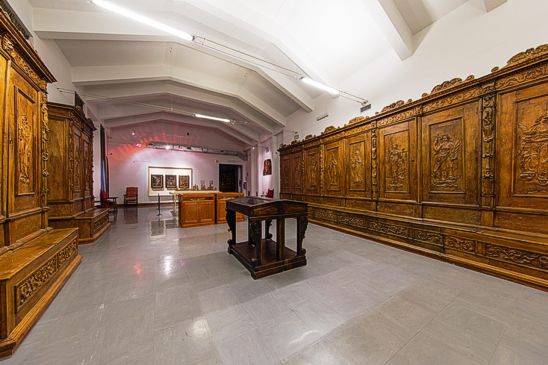 Il Museo Capitolare di Atri, fondato nel 1912 da Mons. Raffaele Tini, è uno dei musei ecclesiastici più antichi d'Italia e rappresenta una testimonianza significativa della storia religiosa e culturale della città. La collezione del museo spazia dal XIII al XX secolo e comprende una vasta gamma di opere d'arte che vanno dalla miniatura alla pittura, dalla scultura all'oreficeria. Il museo è noto per le sue donazioni, che hanno arricchito ulteriormente il patrimonio esposto, tra cui una sala interamente dedicata alla ceramica abruzzese, un settore particolarmente apprezzato per la sua rilevanza artistica e storica. L'edificio che ospita il museo ha una storia che si intreccia con quella della città di Atri. Originariamente costruito come Monastero dei Benedettini nel XII secolo, nel corso dei secoli ha subito diverse trasformazioni. Nel XV secolo divenne la residenza dei canonici, e nel XX secolo ha ospitato il Museo Capitolare. Durante gli anni '60, sotto la supervisione del soprintendente Guglielmo Matthiae, l'edificio fu ristrutturato, con la demolizione e la ricostruzione dell'ala nord, creando uno spazio più razionale per l'esposizione delle opere. Questo intervento ha permesso di esporre in modo ottimale anche gli armadi scolpiti da Carlo Riccione, che sono diventati una delle caratteristiche distintive del museo. Nel corso degli anni, il museo ha continuato a crescere grazie a numerose donazioni, tra cui quelle di ceramiche provenienti da Vincenzo Bindi e opere d'arte lignea da Tommaso Illuminati. All'interno delle sale del museo, i visitatori possono ammirare una ricca collezione di paramenti sacri, tra cui pianete, stole e mitrie del XVII e XVIII secolo, ma anche opere lignee e sculture sacre. La sezione dedicata all'arte tessile sacra è particolarmente significativa, con paramenti realizzati in seta, velluto, broccato e damasco, alcuni dei quali provengono da donazioni storiche, come il tappeto ricamato in argento donato nel 1732 al cardinale Troiano Acquaviva dalla regina d'Inghilterra. Il museo vanta anche una notevole pinacoteca, che espone opere pittoriche di grande valore, come le tavole attribuite a Pedro de Aponte, datate tra il 1500 e il 1507, e altre opere legate alle scuole veneto-friulane e abruzzesi. Tra queste spiccano il Polittico della Madonna col Bambino e i Santi, risalente alla fine del XV secolo, e la Madonna col Bambino in legno scolpito, che rappresenta un esempio pregevole di arte abruzzese del XIII secolo. Oltre alle opere pittoriche, il museo ospita anche una vasta collezione di ceramiche abruzzesi, tra cui pezzi della scuola di Castelli, che illustrano l'evoluzione della ceramica regionale dal XVI al XIX secolo.