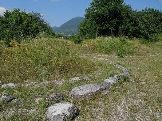 Borghi Medievali e Archeologia in Abruzzo: Campli e la Necropoli di Campovalano. Borghi Medievali in Abruzzo: Campli e la Necropoli di Campovalano. Nel cuore dell’Abruzzo, tra le dolci colline teramane e le pendici dei Monti della Laga, sorge Campli, un borgo medievale dal fascino senza tempo. Le sue stradine acciottolate, le piazze armoniose e gli antichi palazzi raccontano secoli di storia, testimoni di un passato glorioso che ancora oggi si respira in ogni angolo. Passeggiando per il centro storico, si incontrano edifici di grande pregio architettonico, tra cui il Palazzo Farnese, la Scala Santa e le suggestive chiese che custodiscono opere d’arte di inestimabile valore. A pochi chilometri dal borgo, la piana di Campovalano custodisce uno dei più straordinari siti archeologici d’Abruzzo. Tra il XIII e il IV secolo a.C., questa vasta area fu una necropoli di enorme importanza, un luogo di sepoltura che ha restituito migliaia di tombe appartenenti all’aristocrazia locale. Gli scavi archeologici hanno riportato alla luce corredi funerari di straordinaria ricchezza, testimoniando il prestigio e la raffinatezza delle popolazioni italiche che abitarono queste terre. Il Museo Archeologico Nazionale di Campli espone i reperti più significativi provenienti dalla necropoli, offrendo ai visitatori un viaggio affascinante attraverso le antiche civiltà che popolarono l’area. Oltre alla preziosa collezione archeologica, il museo rappresenta un ponte tra passato e presente, permettendo di comprendere l’importanza culturale e storica di questo territorio. Il legame tra il borgo e la necropoli è profondo, un filo invisibile che intreccia Medioevo e antichità in un unico grande racconto. Campli e la Necropoli di Campovalano offrono un’esperienza unica, un viaggio attraverso epoche lontane in uno scenario naturale di straordinaria bellezza. L’atmosfera medievale del borgo e il mistero delle antiche tombe creano un contrasto suggestivo, invitando il visitatore a immergersi in una storia millenaria che continua a vivere tra le pietre, le colline e i silenzi di questo angolo d’Abruzzo.