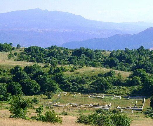 Pallanum fu un’importante città dell’Italia antica, tradizionalmente attribuita al popolo italico dei Lucani, oggi identificata con il suggestivo sito archeologico che sorge sul Monte Pallano, nel territorio di Tornareccio, in provincia di Chieti. Il nome Pallanum compare per la prima volta nella Tavola Peutingeriana, preziosa testimonianza cartografica di età romana, che documenta il ruolo del centro lungo la strada che collegava Aternum, l’attuale Pescara, a Larinum, nei pressi dell’odierna Larino. La città si trovava a circa sette miglia da Anxanum, l’attuale Lanciano, e a dodici miglia da Histonium, oggi Vasto, confermando la sua posizione strategica nel sistema viario dell’Adriatico centrale. Secondo lo storico Edward Bispham, Pallanum avrebbe origini sannitiche, mentre a partire dal I secolo a.C. si registra un progressivo calo della popolazione, preludio al declino del centro. Tra il XVIII e il XIX secolo il sito attirò l’interesse di importanti storiografi, tra cui Pietro Polidori, Domenico Romanelli e Antonio Ludovico Antinori, mentre in epoca contemporanea fu oggetto di studi archeologici da parte di Valerio Cianfarani e Andrea Staffa. Le campagne di scavo condotte negli anni Novanta hanno restituito numerosi reperti in ceramica e pietra, oggi conservati ed esposti a Chieti presso il Convitto Nazionale “Giambattista Vico” e il Museo Archeologico Nazionale d’Abruzzo di Villa Frigerj. Uno degli elementi più spettacolari del sito è rappresentato dalle imponenti mura difensive di età italica, che si sviluppano sul lato orientale dell’antico abitato. Di questa poderosa cinta muraria si conservano circa 160 metri, con un’altezza media di quattro metri, realizzati con grandi blocchi di calcare locale disposti a secco. In origine le mura erano interrotte da quattro porte di accesso, delle quali ne sono sopravvissute due: la Porta del Piano, situata nella parte più elevata del sito e coperta da un architrave, e la Porta del Monte, collocata più a valle, dove i blocchi risultano di dimensioni minori, segno di una diversa tecnica costruttiva. La tradizione popolare attribuisce erroneamente la costruzione delle mura a Carlo Magno, ma le evidenze archeologiche ne collocano la realizzazione tra il VI e il III secolo a.C. A ovest della cinta megalitica, in un avvallamento posto a quota leggermente inferiore, si estendono i resti di un insediamento romano-ellenistico di medie dimensioni, articolato attorno a uno spazio centrale aperto. Qui sono state rinvenute tegole in terracotta, monete di età romana e numerosi manufatti in ferro, mentre le strutture murarie presentano sia tecniche a secco sia l’opera pseudo-isodoma. Le pavimentazioni in opus signinum, realizzate con argilla, gesso e pozzolana, insieme ai marcati dislivelli del terreno, suggeriscono un abitato organizzato su più terrazzamenti, testimoniando la complessità e la lunga continuità di vita del sito di Pallanum.
