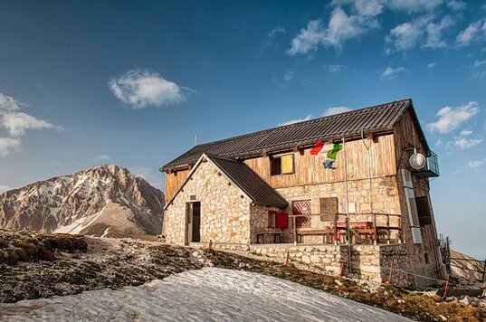 Il Rifugio Duca degli Abruzzi Il Rifugio Duca degli Abruzzi è una delle mete più affascinanti per gli amanti della montagna in Abruzzo. Situato sulla Cresta del Monte Portella, a 2.388 metri di altitudine, offre una vista mozzafiato sulle vette del Gran Sasso e sul vasto altopiano di Campo Imperatore. La sua posizione privilegiata lo rende un punto di riferimento per escursionisti, alpinisti e appassionati di natura che desiderano esplorare questo straordinario angolo dell'Appennino centrale. La struttura, accogliente e immersa in un contesto paesaggistico unico, rappresenta un rifugio ideale per chi cerca una pausa dal caos quotidiano. Durante l'estate, il rifugio è frequentato da escursionisti che affrontano le numerose vie di trekking della zona, mentre in inverno diventa un punto strategico per gli amanti dello sci alpinismo e delle escursioni sulla neve. L'atmosfera familiare e la calorosa ospitalità rendono ogni soggiorno un’esperienza autentica e indimenticabile. Il rifugio non è solo un punto di appoggio per chi esplora il massiccio del Gran Sasso, ma anche un luogo dove assaporare la cucina tipica abruzzese. I piatti serviti sono preparati con ingredienti genuini e locali, offrendo sapori autentici che rispecchiano la tradizione gastronomica della regione. La semplicità della cucina di montagna si combina con il piacere di un pasto caldo dopo una lunga escursione, rendendo il soggiorno ancora più piacevole. Oltre alla gastronomia e alle escursioni, il rifugio rappresenta un luogo di condivisione per gli amanti della montagna. Le serate trascorse davanti al camino, con racconti di avventure ed esperienze vissute in alta quota, creano un’atmosfera speciale di convivialità e amicizia. Lontano dalle luci artificiali delle città, il cielo stellato sopra il rifugio regala spettacoli indimenticabili, permettendo di ammirare la bellezza della natura incontaminata. Il Rifugio Duca degli Abruzzi è molto più di una semplice meta per escursionisti; è un luogo che permette di riconnettersi con la montagna e con sé stessi. Immerso in un paesaggio selvaggio e suggestivo, offre la possibilità di vivere un’esperienza unica, fatta di avventura, relax e contatto con la natura. Chiunque vi giunga porta con sé un ricordo indelebile, fatto di emozioni, panorami spettacolari e il calore di un’accoglienza autentica.