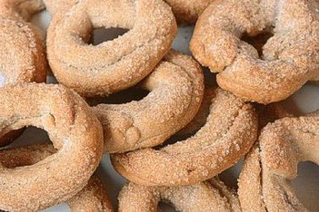 Per preparare i tarallucci al vino, iniziate mescolando la farina con il lievito e un pizzico di sale in una ciotola capiente. Aggiungere poi lo zucchero, che darà al dolce una leggera dolcezza, e mescolate bene gli ingredienti secchi. In un'altra ciotola, unite l'olio di semi e il vino bianco, che daranno morbidezza all'impasto e un aroma particolare. Incorporate gradualmente gli ingredienti liquidi a quelli secchi, mescolando con le mani fino a ottenere un impasto omogeneo e morbido. Se necessario, aggiungete un po' di farina per ottenere la giusta consistenza, che deve essere facilmente lavorabile. Una volta che l'impasto è pronto, prelevate delle piccole porzioni e formate dei cilindri di pasta, che dovranno essere lunghi circa 5 cm. Ungete leggermente le mani per facilitare la lavorazione. Chiudete ogni cilindro di pasta a formare un anello, sigillando bene le estremità. Disponete i tarallucci su una teglia rivestita di carta forno, facendo attenzione a lasciare un po' di spazio tra un taralluccio e l'altro, in modo che non si attacchino durante la cottura. Prima di infornare, se lo desiderate, potete spolverizzare i tarallucci con dello zucchero semolato, che durante la cottura conferirà loro una dolce crosticina. Cuocete i tarallucci in forno preriscaldato a 180°C per circa 15-20 minuti, o finché non saranno ben dorati e croccanti. Durante la cottura, l’impasto crescerà leggermente, formando delle piccole ciambelline fragranti e dorate. Una volta pronti, togliete i tarallucci dal forno e lasciateli raffreddare su una griglia. Dopo che si sono completamente raffreddati, saranno pronti per essere serviti. I tarallucci al vino sono perfetti per essere accompagnati da un buon tè o un caffè, e rappresentano un dolce semplice e gustoso della tradizione abruzzese, particolarmente apprezzato per la sua leggerezza e il suo sapore delicato.