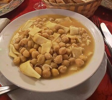 Per preparare le Taccozze e ceci, si inizia con la preparazione della pasta. La semola di grano duro, un prodotto tipico abruzzese, viene mescolata con un uovo e un pizzico di sale. L'impasto, lavorato a mano fino a ottenere una consistenza elastica e liscia, viene poi steso su una superficie infarinata e tagliato a strisce larghe, creando le classiche taccozze. Questa pasta ruvida è perfetta per assorbire il brodo ricco dei ceci, un legume che è un altro prodotto tradizionale della regione. Dopo aver lasciato riposare l'impasto, si cuociono le taccozze in acqua salata bollente, fino a che non risultano al dente. Nel frattempo, si prepara il sugo di ceci. I ceci, che devono essere messi in ammollo per una notte prima di essere utilizzati, sono cucinati lentamente in acqua con cipolla e aglio, due ingredienti fondamentali per insaporire il brodo. L'aggiunta di un rametto di rosmarino, erba aromatica tipica dell'Abruzzo, conferisce al piatto una fragranza naturale che esalta il sapore dei legumi. Il peperoncino, che non può mancare nella cucina abruzzese, dona al sugo una leggera piccantezza, che si bilancia perfettamente con la dolcezza dei ceci. Quando i ceci sono ben cotti e il brodo ha acquisito una consistenza densa e saporita, si aggiungono le taccozze cotte, mescolando il tutto per far sì che la pasta assorba il sapore del brodo. In questo piatto, il brodo diventa un vero protagonista, avvolgendo la pasta con il suo sapore ricco e confortante. La combinazione dei ceci, con la loro consistenza cremosa, e delle taccozze, che assorbono il brodo, crea un equilibrio perfetto di consistenze e sapori. Per completare il piatto, si aggiunge un filo d'olio extravergine d'oliva, un altro prodotto tipico abruzzese che aggiunge un tocco finale di sapore e cremosità. L'olio, con il suo gusto fruttato e leggermente piccante, esalta ulteriormente la ricchezza del brodo e rende il piatto ancora più avvolgente. Le Taccozze e ceci sono un piatto che rappresenta la tradizione della cucina povera e genuina della provincia di Pescara, fatto di ingredienti semplici ma dal sapore deciso e autentico.