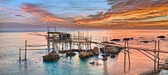 Il mare e la montagna in Abruzzo: La costa dei Trabocchi. La Costa dei Trabocchi, situata lungo il litorale meridionale dell'Abruzzo, è una delle meraviglie più affascinanti della regione. Questo tratto costiero si distingue per la presenza dei suggestivi trabocchi, antiche macchine da pesca interamente costruite in legno. Sorrette da lunghi pali infissi nel fondale marino, queste strutture si protendono verso il mare, collegate alla terraferma da passerelle anch'esse di legno. La loro immagine, soprattutto al tramonto o durante le mareggiate, restituisce un'atmosfera unica e senza tempo, testimoniando un passato legato alla tradizione marinara. Lungo la costa, il paesaggio cambia in modo spettacolare. Dalle ampie spiagge sabbiose si passa a tratti rocciosi e frastagliati, con falesie che si tuffano nelle acque cristalline dell'Adriatico. Questa varietà morfologica ha favorito la conservazione di un ambiente incontaminato, dove la vegetazione mediterranea si mescola con il profumo della salsedine. Ogni curva della strada costiera offre scorci incantevoli, con vedute che si aprono su calette nascoste e piccoli borghi di pescatori, custodi di antiche usanze e sapori autentici. Uno dei luoghi più evocativi della Costa dei Trabocchi è Fossacesia, con il suo celebre trabocco di Capo Turchino, immortalato dal poeta Gabriele d'Annunzio nelle sue opere. Qui, tra il verde degli uliveti e l'azzurro del mare, sorge l'Abbazia di San Giovanni in Venere, un imponente monastero benedettino che domina il paesaggio con la sua maestosa architettura. Non lontano, il promontorio dannunziano regala panorami indimenticabili, dove la natura e la storia si fondono in un'armonia perfetta. Proseguendo verso sud, si incontrano altre meraviglie come il trabocco di Punta del Cavalluccio e la spiaggia di Vallevò, angoli di paradiso in cui il tempo sembra essersi fermato. Qui, il mare limpido lambisce le rive di ciottoli, mentre le strutture in legno dei trabocchi si stagliano contro l'orizzonte, creando un quadro di straordinaria bellezza. La Costa dei Trabocchi rappresenta un'esperienza unica, un viaggio tra storia, natura e tradizione che lascia nel cuore di chi la visita un ricordo indelebile.